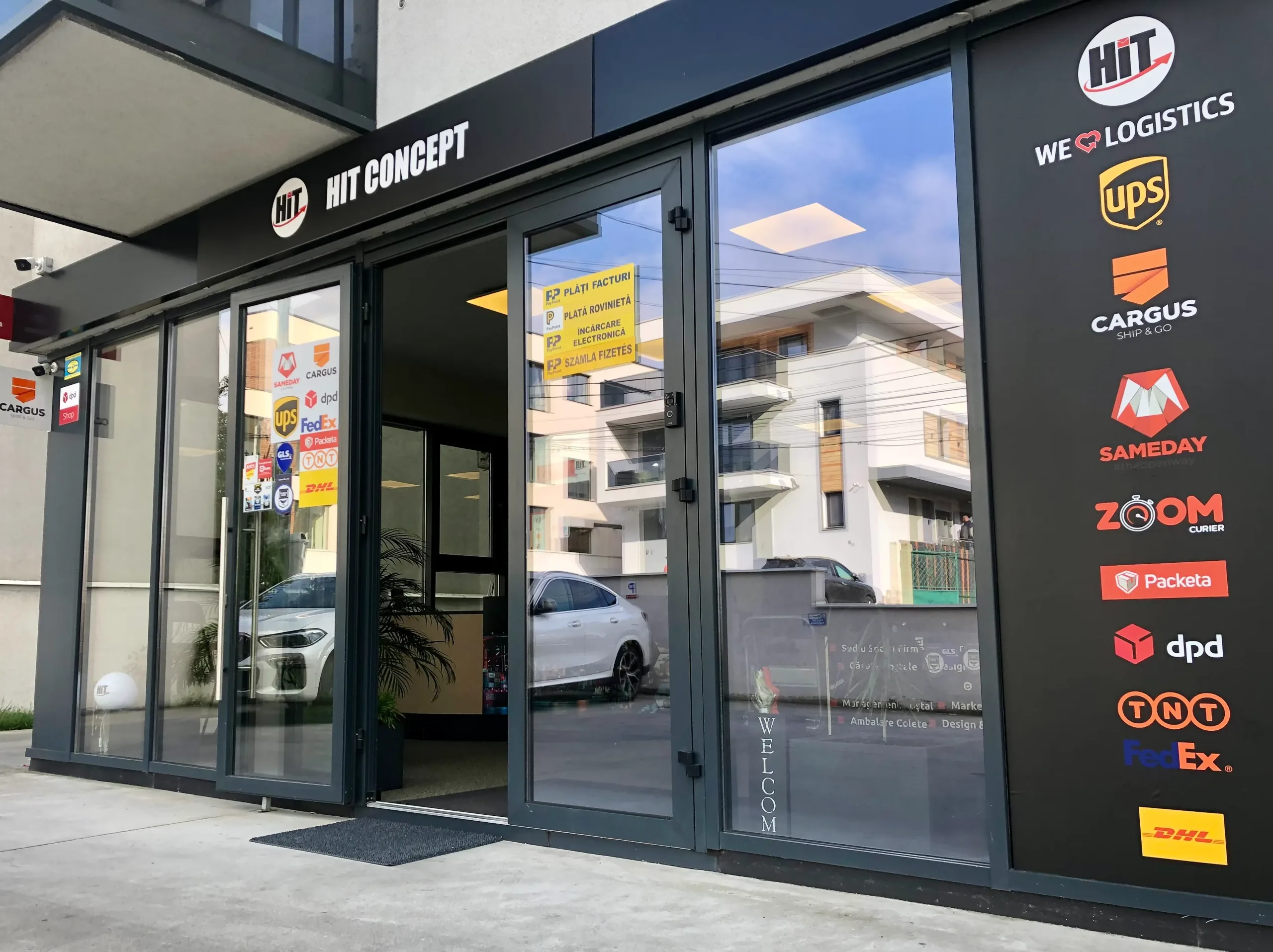 Vedere exterioară a magazinului de logistică HIT CONCEPT cu siglele principalelor companii de transport aerian, maritim și curierat.